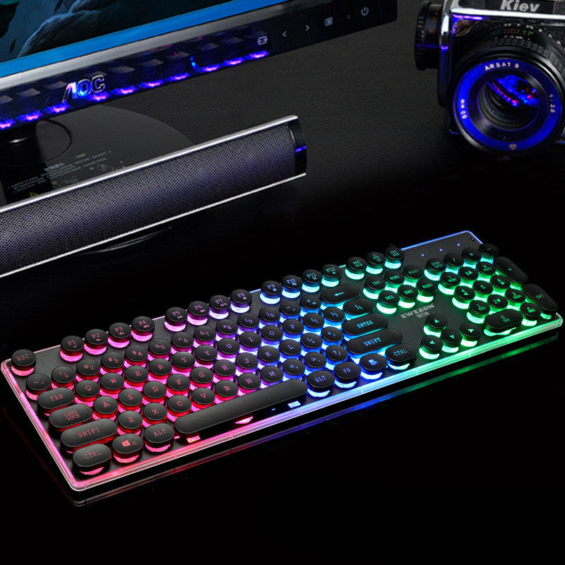 Gaming keyboard with RGB lighting on a dark surface with computer monitor and speakers.