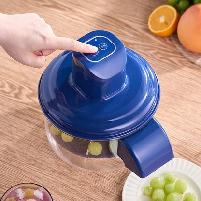 Blue manual juicer with a hand pressing a button, surrounded by fruits on a wooden table.