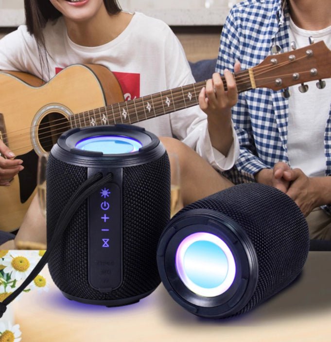 Two black cylindrical speakers with colorful lights on a table, with people playing guitar in the background.