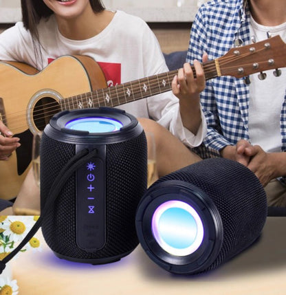 Two black cylindrical speakers with colorful lights on a table, with people playing guitar in the background.