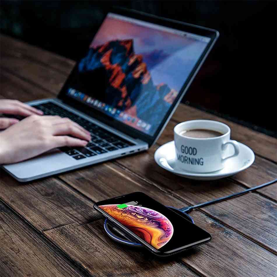Person using a laptop with a smartphone on a wireless charger, next to a cup of coffee on a wooden table.