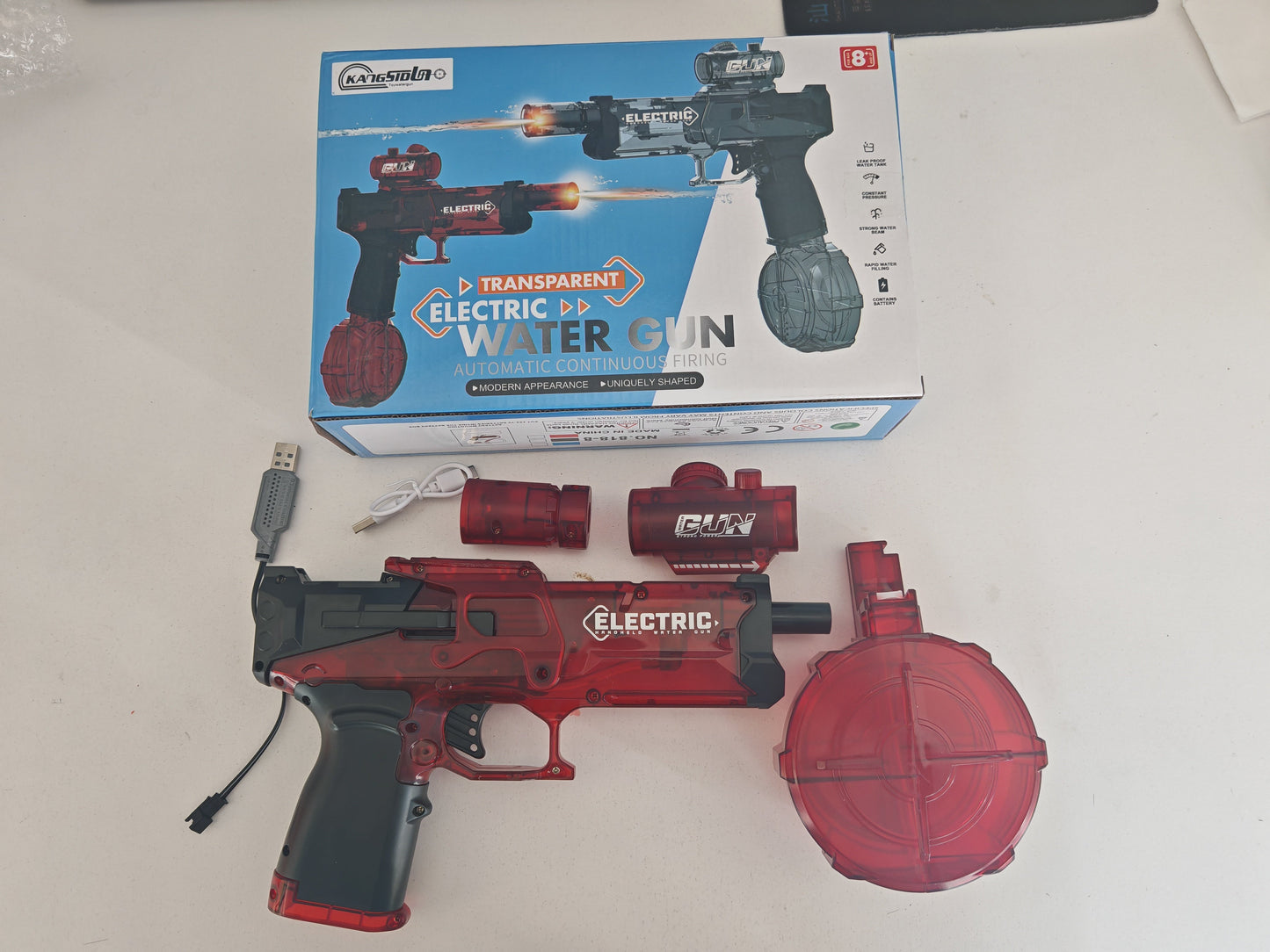 Red electric water gun with USB cable and transparent water tank on a white surface, with packaging in the background.
