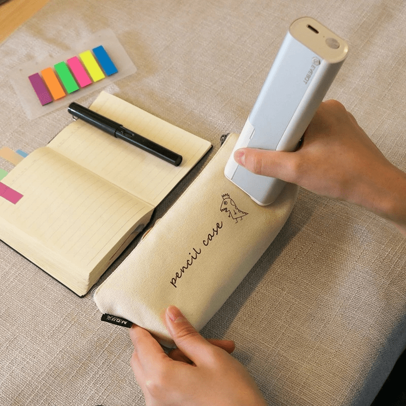 Hand holding a small printer over a pencil case with a notebook and colorful markers on a textured surface.