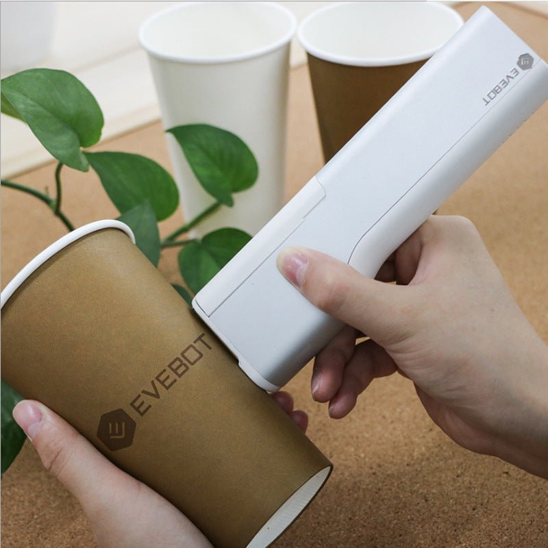 Hand using a white portable printer device labeled 'Evebot' on a brown cylindrical container with green leaves in the background.
