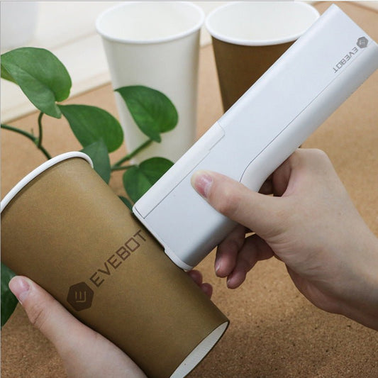 Hand using a white portable printer device labeled 'Evebot' on a brown cylindrical container with green leaves in the background.