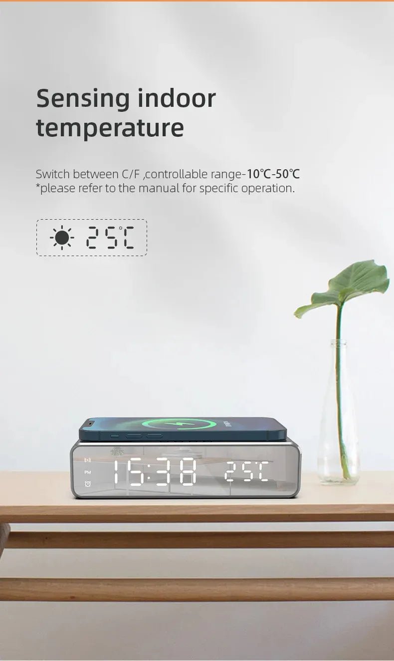Digital clock with temperature display on a wooden surface, featuring a plant in the background.
