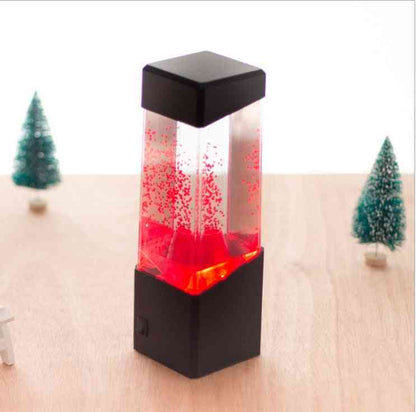 Lava lamp with red liquid and black base on a light wooden surface with small Christmas trees in the background.