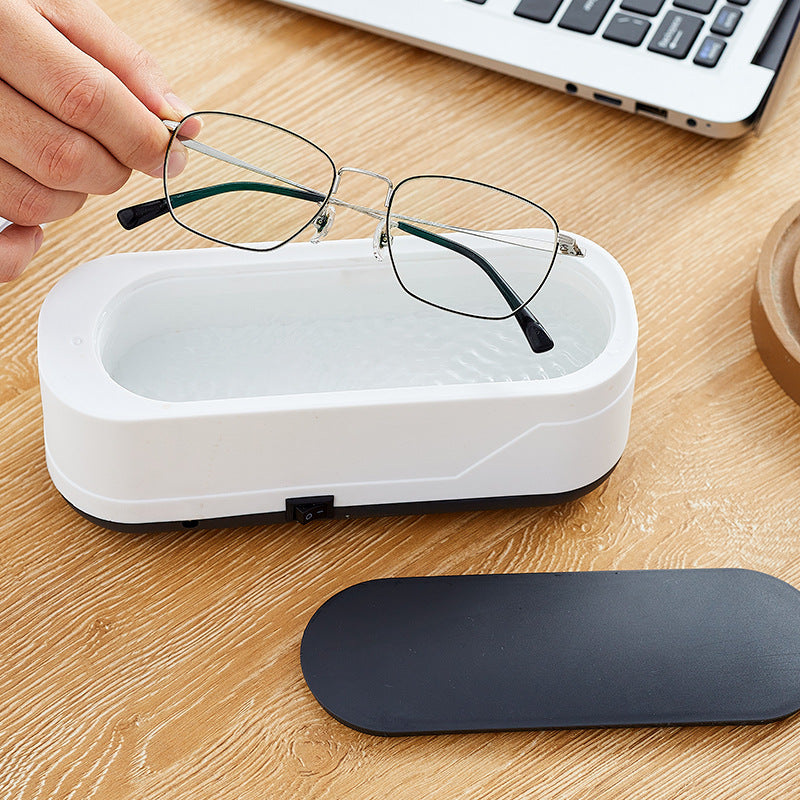 Person placing glasses into a white cleaning case on a wooden desk with a laptop in the background.