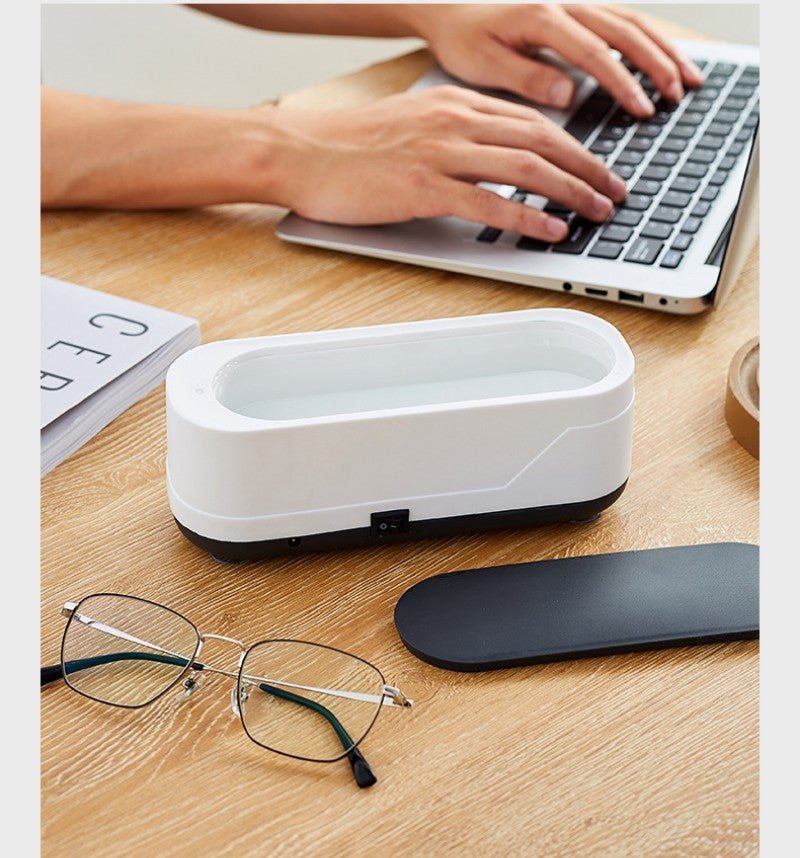 White electronic device on a desk with glasses and a mouse nearby, person using a laptop in the background.