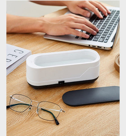 White electronic device on a desk with glasses and a mouse nearby, person using a laptop in the background.