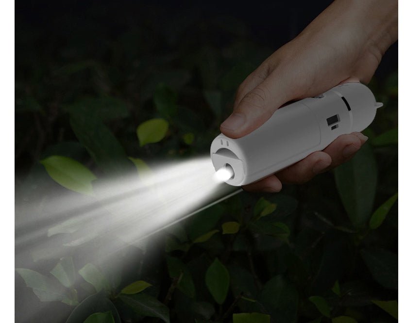 Hand holding a white flashlight emitting light in a dark outdoor setting with greenery.