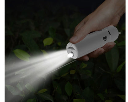 Hand holding a white flashlight emitting light in a dark outdoor setting with greenery.